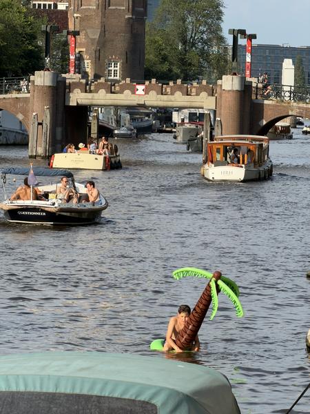 This vibrant photo captures a lively scene on the canals of Oudeschans, Uilenburg, Amsterdam, with several boats carrying people enjoying the sunny weather